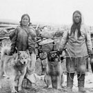 Photo d'un homme et d'une femme inuits debout devant un mur de pierres avec trois chiens, Fullerton, Nunavut, 1904-1905