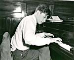 Photograph of Glenn Gould, seated with his legs crossed, playing the piano