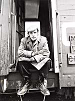 Photograph of Glenn Gould, sitting in the doorway of a caboose, and wearing a cap, overcoat and galoshes